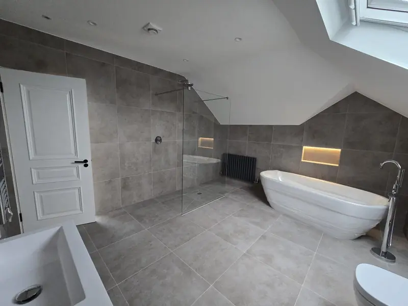 Large grey-tiled bathroom with a freestanding bath and floor-standing tap beneath a sloping ceiling